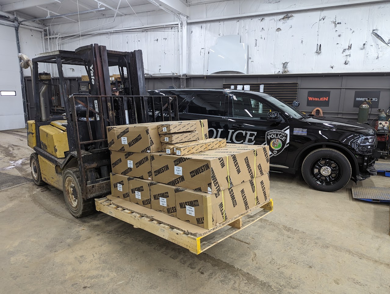 Forklift moving a crated pallet past a police SUV inside a shop